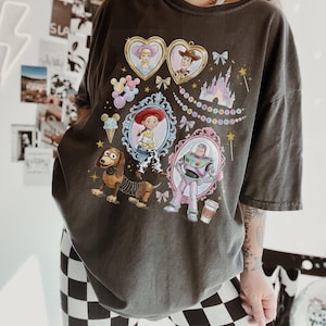 May include: Dark grey t-shirt with a colourful graphic of Toy Story characters in ornate frames. The design includes images of Woody, Buzz Lightyear, and other characters, with the phrase "To infinity and beyond".