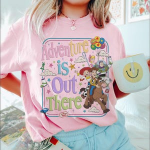Pode incluir: Camiseta rosa claro com a frase "Adventure is Out There" em um design retr&ocirc; colorido. O gr&aacute;fico apresenta personagens de desenhos animados e um logotipo da Pizza Planet. Uma caneca branca com um rosto sorridente amarelo &eacute; segurada.