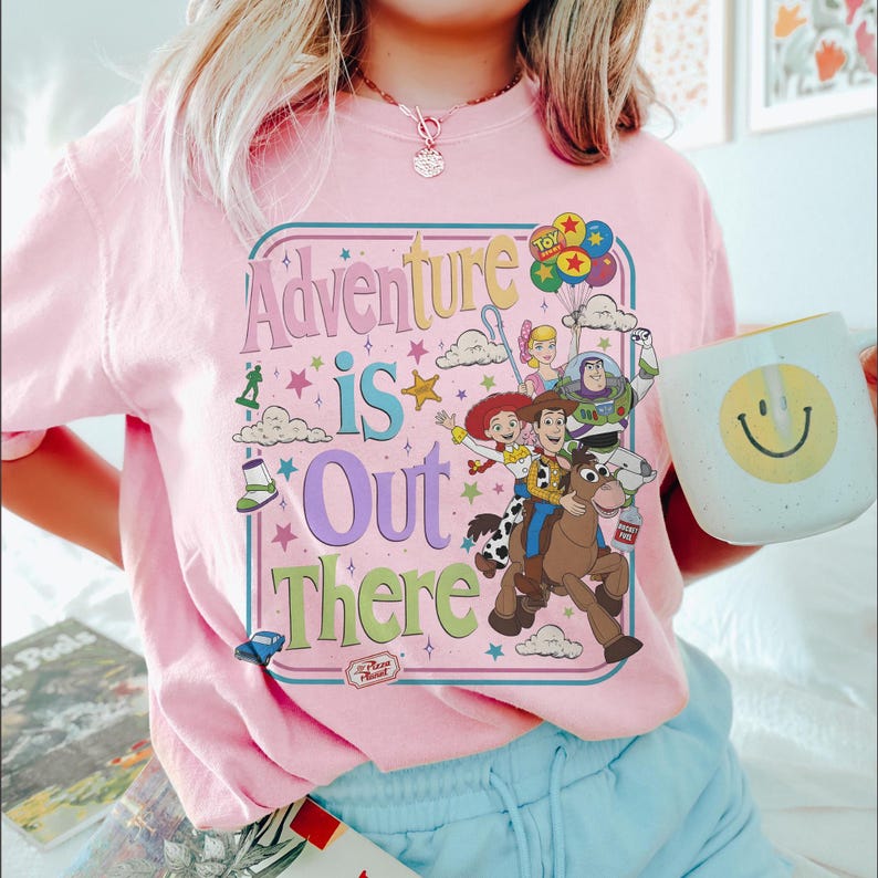 Pode incluir: Camiseta rosa claro com a frase "Adventure is Out There" em um design retr&ocirc; colorido. O gr&aacute;fico apresenta personagens de desenhos animados e um logotipo da Pizza Planet. Uma caneca branca com um rosto sorridente amarelo &eacute; segurada.