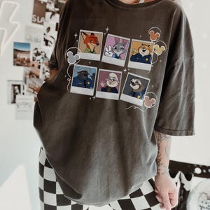 May include: Dark gray t-shirt featuring a collage of cartoon character portraits. The portraits include a fox, a rabbit, a tiger, a buffalo, a sheep, and a sloth. Mickey Mouse-shaped balloons and stars decorate the design.