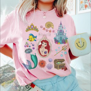Puede incluir: Camiseta rosa claro con ilustraciones coloridas de una pel&iacute;cula animada popular. El dise&ntilde;o incluye una sirena, un castillo, un cofre del tesoro y otros personajes y objetos. En la imagen se sostiene una taza blanca con una carita sonriente amarilla.