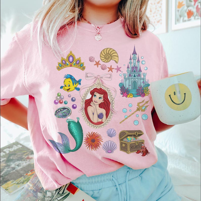 Puede incluir: Camiseta rosa claro con ilustraciones coloridas de una pel&iacute;cula animada popular. El dise&ntilde;o incluye una sirena, un castillo, un cofre del tesoro y otros personajes y objetos. En la imagen se sostiene una taza blanca con una carita sonriente amarilla.