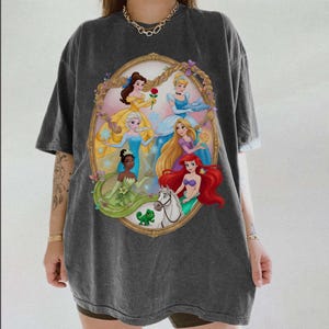 May include: Dark grey t-shirt with a colourful illustration of Disney princesses in a gold oval frame. The princesses are shown in their signature gowns, with details such as a rose and a frog.