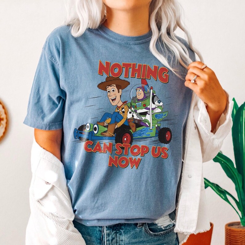 May include: A blue t-shirt featuring a graphic of Woody, Buzz Lightyear, and other characters from Toy Story in a race car. The text "NOTHING CAN STOP US NOW" is printed in red and white.