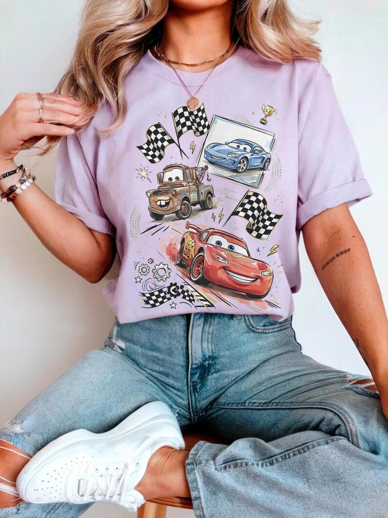 May include: A light purple t-shirt featuring a graphic of animated race cars, including Lightning McQueen, Mater, and a blue car. The design includes checkered flags, a trophy, and a framed image of a car. The shirt is made of a soft material.