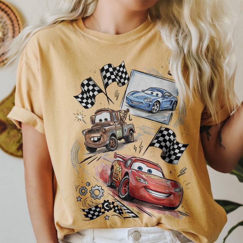 May include: Mustard yellow t-shirt featuring a graphic of animated race cars, including a red car with the number 95, a brown tow truck, and a blue sports car. Checkered flags and lightning bolts are also included in the design.