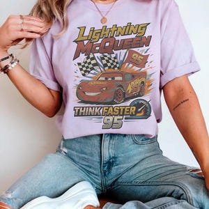 Pode incluir: Uma camiseta lil&aacute;s com um gr&aacute;fico de Rel&acirc;mpago McQueen do filme Carros. O design inclui o texto "Lightning McQueen", "95" e "Think Faster". A camisa &eacute; usada com jeans azul.