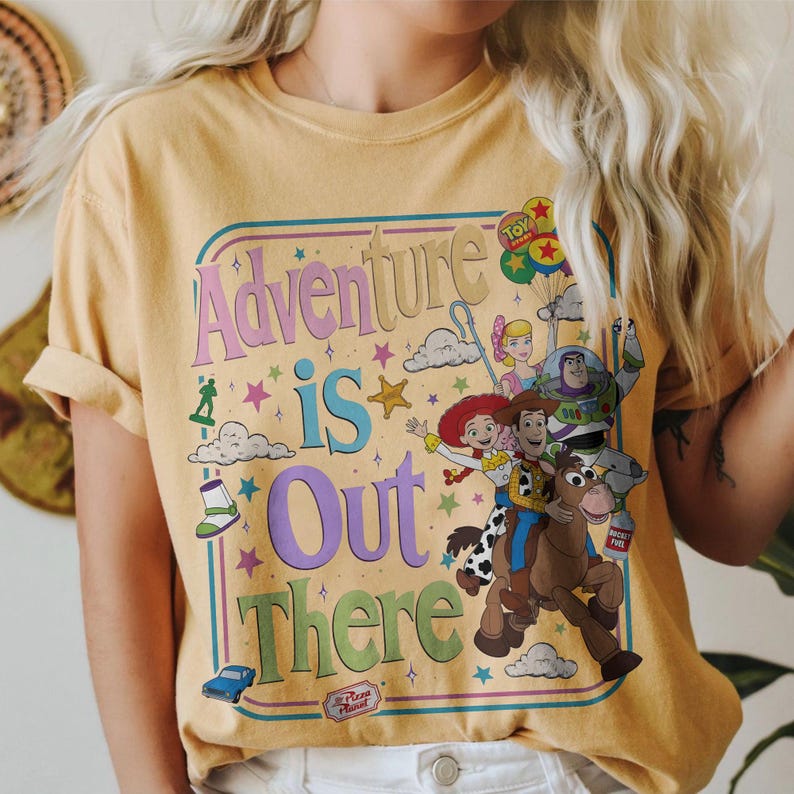 Pode incluir: Camiseta amarelo mostarda com a frase "Adventure is Out There" e personagens de Toy Story. O design inclui Woody, Buzz Lightyear, Jessie e outros personagens. A camiseta tem gola redonda e mangas curtas.