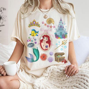 Puede incluir: Camiseta color crema con un collage de ilustraciones tem&aacute;ticas de La Sirenita. Incluye un retrato de Ariel, un castillo, Flounder, una cola de sirena, un cofre del tesoro y otros elementos acu&aacute;ticos.