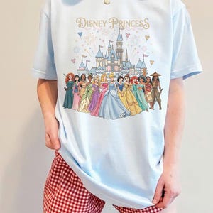 K&ouml;nnte beinhalten: Hellblaues T-Shirt mit dem Schriftzug "Disney Princess" und einer Illustration von Disney-Prinzessinnen vor einem Schloss. Das Shirt wird mit rot-wei&szlig; karierten Shorts getragen.