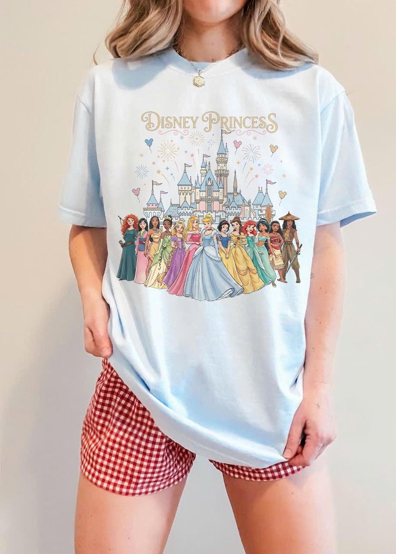 K&ouml;nnte beinhalten: Hellblaues T-Shirt mit dem Schriftzug "Disney Princess" und einer Illustration von Disney-Prinzessinnen vor einem Schloss. Das Shirt wird mit rot-wei&szlig; karierten Shorts getragen.
