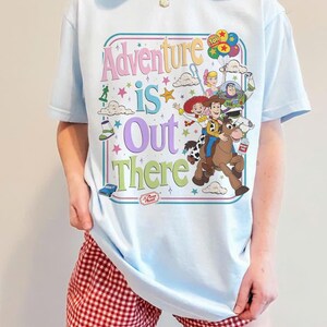 Pode incluir: T-shirt azul claro com um gr&aacute;fico colorido dos personagens de Toy Story e a frase "Adventure is Out There". O design inclui Woody, Buzz Lightyear e outros personagens. A camiseta tem gola redonda.