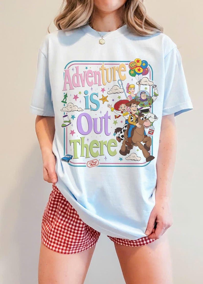 Pode incluir: T-shirt azul claro com um gr&aacute;fico colorido dos personagens de Toy Story e a frase "Adventure is Out There". O design inclui Woody, Buzz Lightyear e outros personagens. A camiseta tem gola redonda.