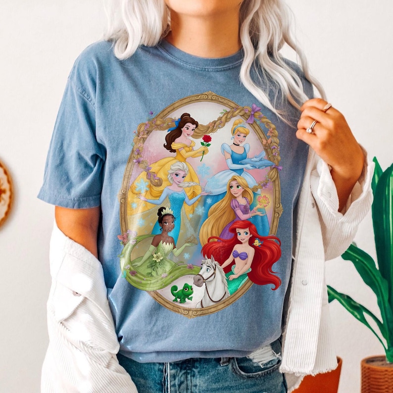 May include: A light blue t-shirt with a circular graphic of animated princesses in a gold frame. The shirt is made of a soft material. The princesses are wearing colorful dresses.