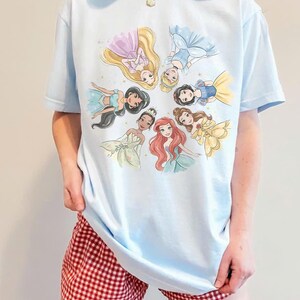 May include: Light blue t-shirt featuring a circular illustration of Disney princesses. The princesses are depicted in their iconic dresses and hairstyles. The shirt is paired with red and white checkered shorts.