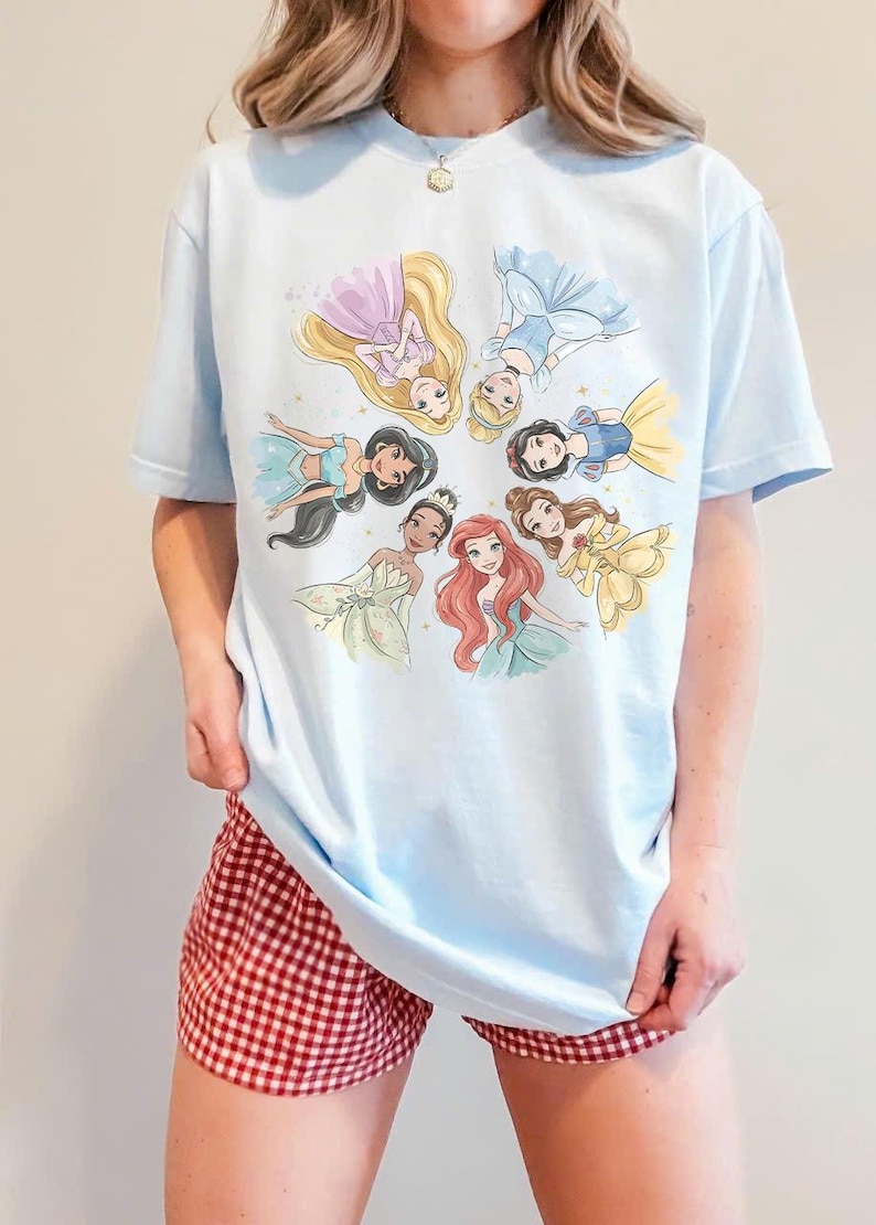 May include: Light blue t-shirt featuring a circular illustration of Disney princesses. The princesses are depicted in their iconic dresses and hairstyles. The shirt is paired with red and white checkered shorts.