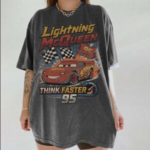 Pode incluir: Camiseta cinza escuro com um design de Rel&acirc;mpago McQueen do filme Carros. O gr&aacute;fico inclui o texto "Lightning McQueen 95" e "Think Faster 95", um carro de corrida vermelho e bandeiras quadriculadas. A camiseta tem um visual vintage.