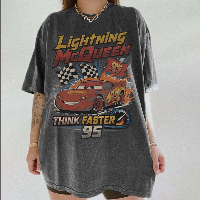 Pode incluir: Camiseta cinza escuro com um design de Rel&acirc;mpago McQueen do filme Carros. O gr&aacute;fico inclui o texto "Lightning McQueen 95" e "Think Faster 95", um carro de corrida vermelho e bandeiras quadriculadas. A camiseta tem um visual vintage.