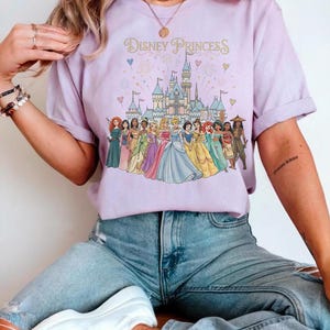 K&ouml;nnte beinhalten: Ein helllila T-Shirt mit dem Text "Disney Princess" &uuml;ber einer Grafik eines Schlosses und mehrerer Disney-Prinzessinnen. Das Shirt wird mit hellblauen Jeans kombiniert.