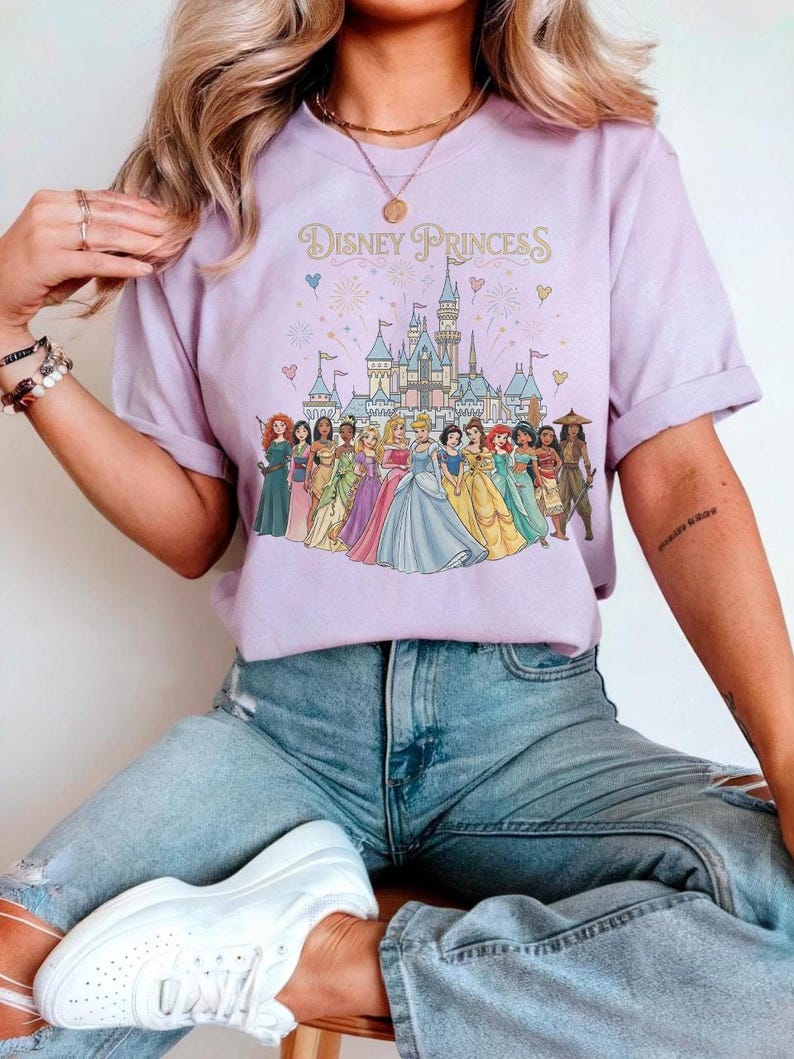 K&ouml;nnte beinhalten: Ein helllila T-Shirt mit dem Text "Disney Princess" &uuml;ber einer Grafik eines Schlosses und mehrerer Disney-Prinzessinnen. Das Shirt wird mit hellblauen Jeans kombiniert.