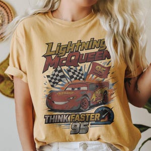 Pode incluir: Camiseta amarelo mostarda com um gr&aacute;fico de Rel&acirc;mpago McQueen do filme Carros. O design inclui o texto "Lightning McQueen", "Think Faster" e o n&uacute;mero "95", juntamente com bandeiras quadriculadas.