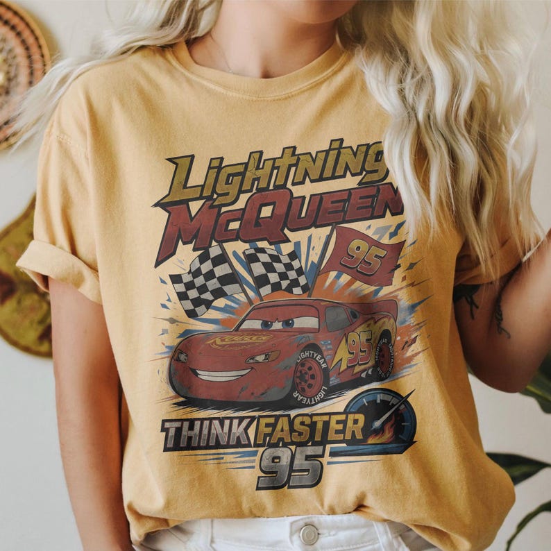 Pode incluir: Camiseta amarelo mostarda com um gr&aacute;fico de Rel&acirc;mpago McQueen do filme Carros. O design inclui o texto "Lightning McQueen", "Think Faster" e o n&uacute;mero "95", juntamente com bandeiras quadriculadas.