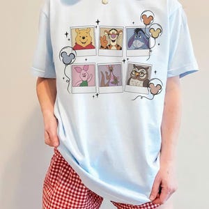 May include: Light blue t-shirt featuring a collage of Winnie the Pooh characters in polaroid-style frames. The shirt has a crew neck and short sleeves. The design includes Winnie the Pooh, Tigger, Eeyore, Piglet, Kanga, Owl, and Mickey Mouse balloons.
