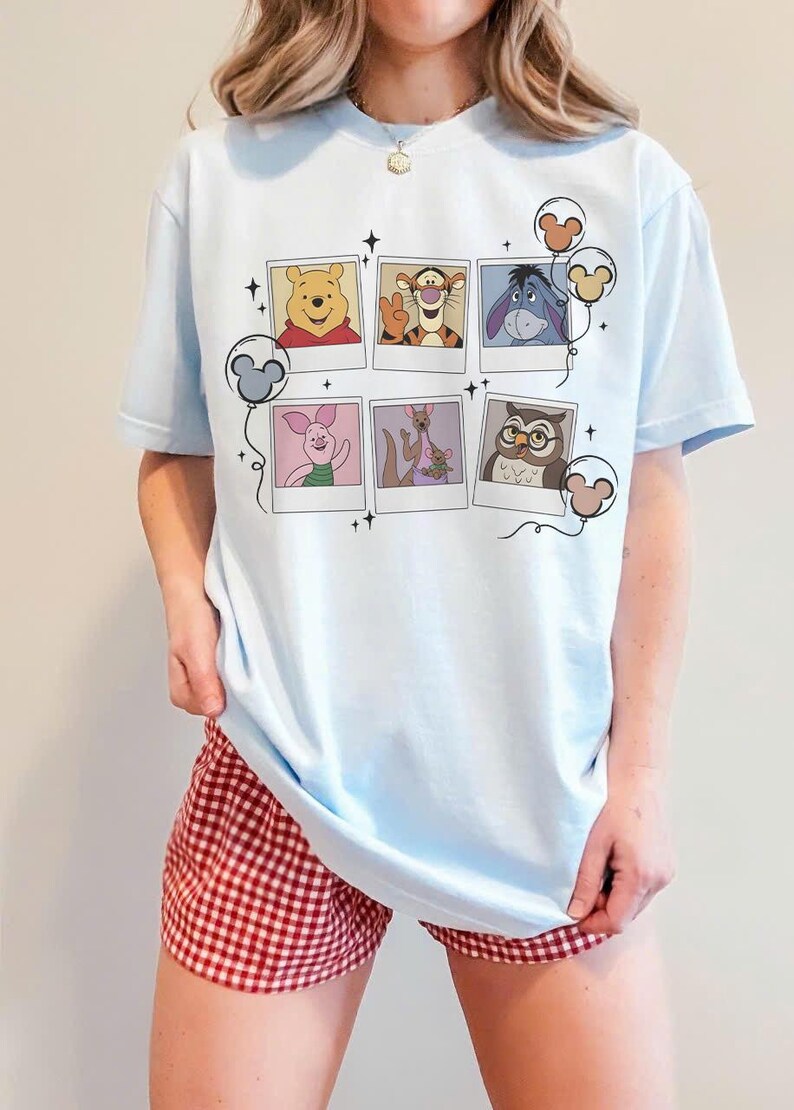 May include: Light blue t-shirt featuring a collage of Winnie the Pooh characters in polaroid-style frames. The shirt has a crew neck and short sleeves. The design includes Winnie the Pooh, Tigger, Eeyore, Piglet, Kanga, Owl, and Mickey Mouse balloons.