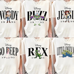 May include: Cream-colored t-shirts featuring Disney's Toy Story characters: Woody, Buzz, Jessie, Bo Peep, Rex, and the Little Green Men. Each shirt has the character's name in bold black letters, the Disney logo, and the year 1995 or 1999.