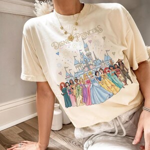 K&ouml;nnte beinhalten: Cremefarbenes T-Shirt mit dem Schriftzug "Disney Princess" &uuml;ber einer farbenfrohen Grafik eines Schlosses und mehrerer Disney-Prinzessinnen. Das Shirt hat kurze &Auml;rmel und eine lockere Passform.