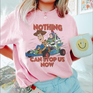 May include: Pink t-shirt featuring a graphic of Woody and Buzz Lightyear from Toy Story in a race car, with the text "NOTHING CAN STOP US NOW". A white mug with a yellow smiley face is held in the hand.
