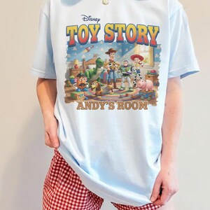 Puede incluir: Camiseta azul claro con el logotipo de Disney Toy Story y personajes. El gr&aacute;fico incluye a Woody, Buzz Lightyear y otros personajes en la habitaci&oacute;n de Andy. La camiseta se combina con pantalones cortos a cuadros rojos y blancos.
