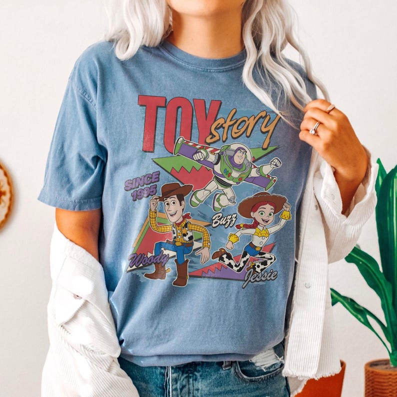 May include: A light blue t-shirt featuring the words "Toy Story" in red and yellow, with the phrase "Since 1995." The shirt displays cartoon images of Woody, Buzz Lightyear, and Jessie. The shirt is a crew neck.