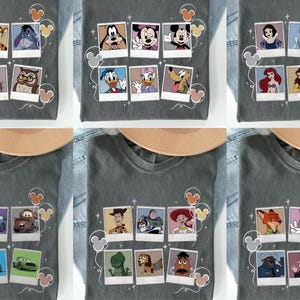 May include: Gray t-shirts featuring various Disney character designs. Each shirt showcases a collection of characters in a polaroid-style arrangement, with Mickey Mouse ears and decorative elements. The shirts are displayed on a denim background.