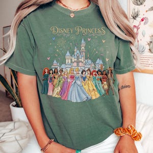 K&ouml;nnte beinhalten: Gr&uuml;nes T-Shirt mit dem Schriftzug "Disney Princess" und einer Grafik eines Schlosses und Disney-Prinzessinnen in bunten Kleidern. Das T-Shirt ist ein l&auml;ssiger Stil, der f&uuml;r den Alltag geeignet ist.