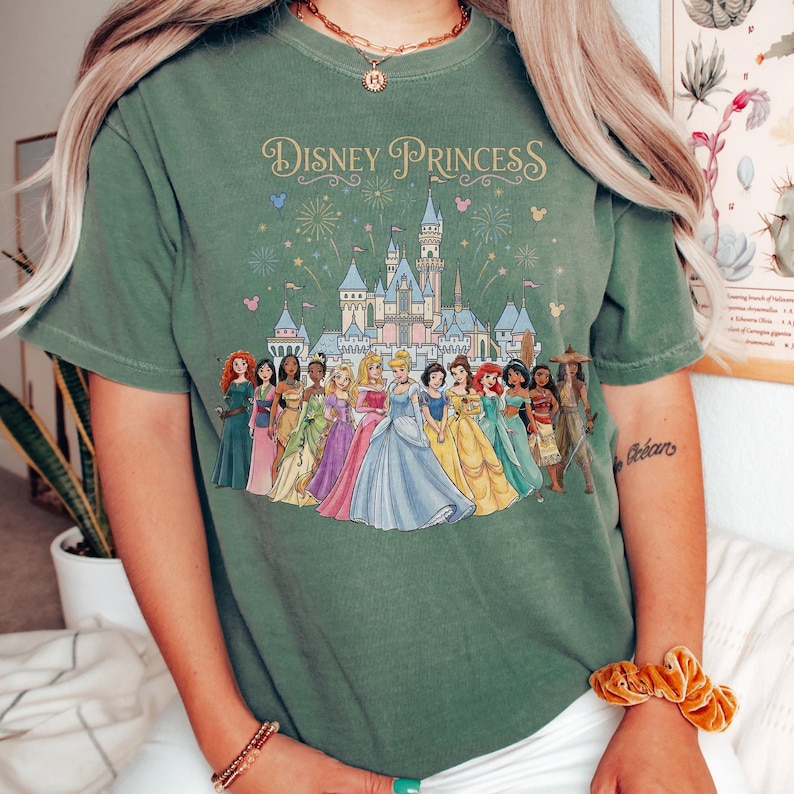 K&ouml;nnte beinhalten: Gr&uuml;nes T-Shirt mit dem Schriftzug "Disney Princess" und einer Grafik eines Schlosses und Disney-Prinzessinnen in bunten Kleidern. Das T-Shirt ist ein l&auml;ssiger Stil, der f&uuml;r den Alltag geeignet ist.