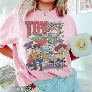 May include: Pink t-shirt featuring the words "Toy Story" above illustrations of Woody, Buzz Lightyear, and Jessie. The shirt also includes the text "Since 1995". A white mug with a yellow smiley face is held in the image.