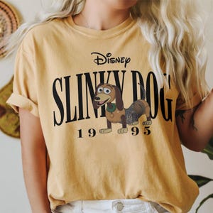 May include: Mustard yellow t-shirt featuring the Disney logo and the words "Slinky Dog" above an illustration of the Slinky Dog character. The number "1995" is printed below the illustration. The shirt has short sleeves.