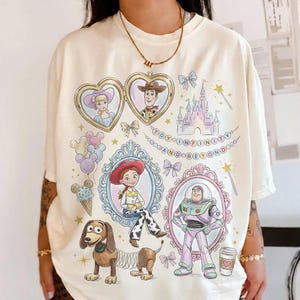 May include: Cream-coloured t-shirt featuring cartoon illustrations of characters from the movie Toy Story. The design includes portraits of Woody, Buzz Lightyear, Jessie, and others, along with the text "TO INFINITY AND BEYOND".