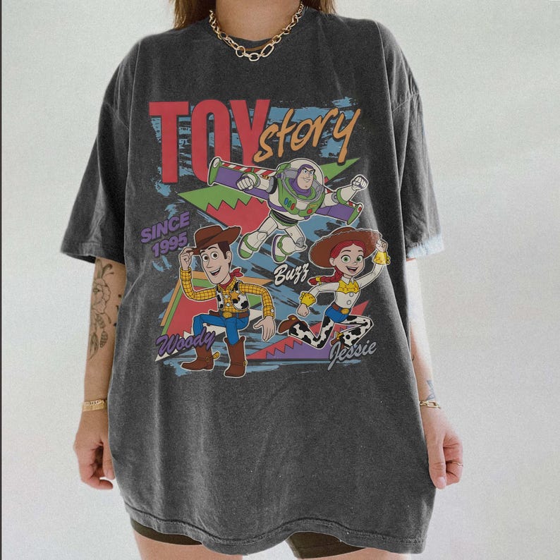 May include: Dark gray t-shirt featuring the Toy Story logo and characters Woody, Buzz, and Jessie. The shirt has the text "Since 1995" and a distressed vintage look. A fun, casual tee for fans of the animated movie.