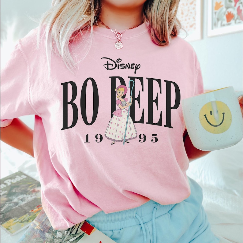 May include: A light pink t-shirt featuring the Disney logo and the words "Bo Peep" in large black letters. Below the text is an illustration of Bo Peep and the numbers "1995". A white mug with a yellow smiley face is held in the hand.