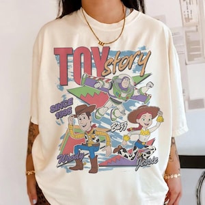 May include: Cream-colored t-shirt featuring a graphic of Toy Story characters Woody, Buzz Lightyear, and Jessie. The design includes the text "Toy Story" and "Since 1995" in a retro style. The shirt has short sleeves.