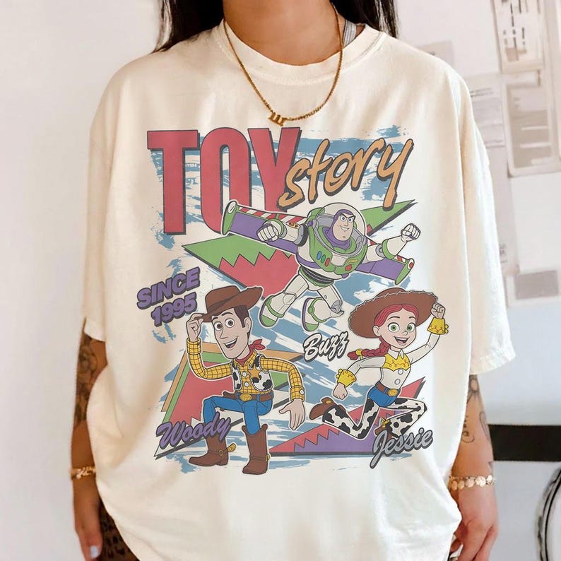 May include: Cream-colored t-shirt featuring a graphic of Toy Story characters Woody, Buzz Lightyear, and Jessie. The design includes the text "Toy Story" and "Since 1995" in a retro style. The shirt has short sleeves.