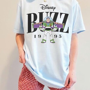 May include: Light blue t-shirt featuring a graphic of Buzz Lightyear from Disney's Toy Story. The design includes the word "Buzz", the Disney logo, and the year "1995". The shirt is a casual wear item.