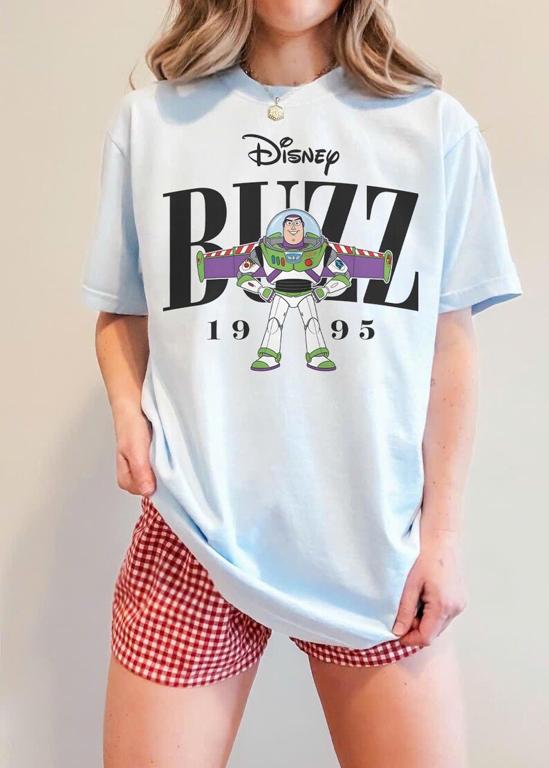 May include: Light blue t-shirt featuring a graphic of Buzz Lightyear from Disney's Toy Story. The design includes the word "Buzz", the Disney logo, and the year "1995". The shirt is a casual wear item.
