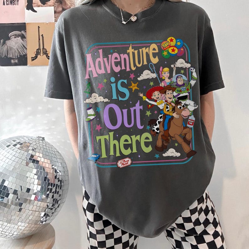Pode incluir: T-shirt cinza escuro com um gr&aacute;fico colorido do filme Toy Story. O design inclui a frase "Adventure is Out There" com imagens de Woody, Buzz Lightyear e outros personagens. A camisa tem mangas curtas.