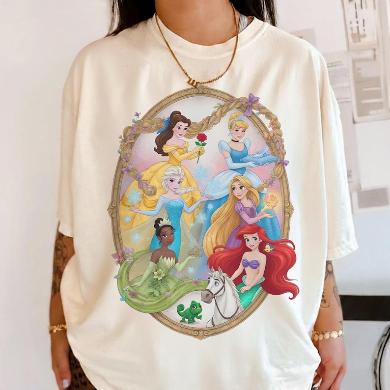 May include: Cream-coloured t-shirt with a colourful illustration of Disney princesses in an oval frame. The princesses are shown in their iconic gowns, with floral and fairytale elements.