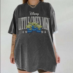 May include: Dark gray t-shirt featuring the Disney logo and the text "Little Green Men 1995." The shirt has a graphic of three green aliens wearing blue uniforms. The shirt is oversized and has short sleeves.