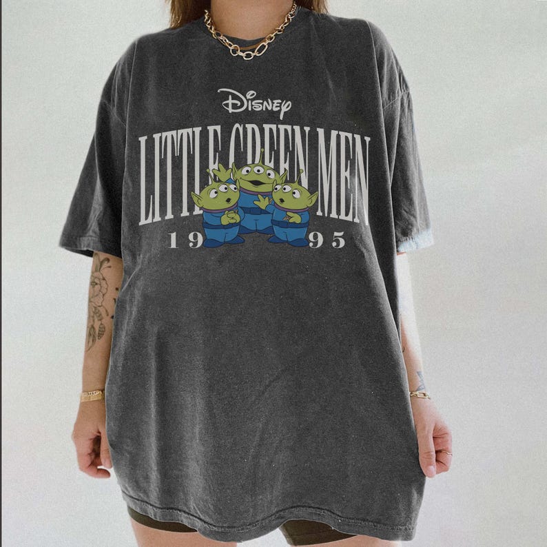 May include: Dark gray t-shirt featuring the Disney logo and the text "Little Green Men 1995." The shirt has a graphic of three green aliens wearing blue uniforms. The shirt is oversized and has short sleeves.