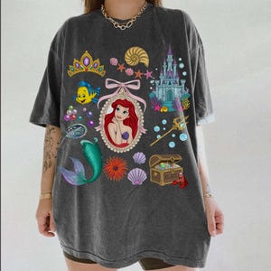 Puede incluir: Camiseta gris oscuro con un gr&aacute;fico colorido de Ariel de La Sirenita, una cola de sirena, un castillo y otras ilustraciones con tem&aacute;tica marina. El dise&ntilde;o incluye una tiara, conchas marinas y un cofre del tesoro. La camiseta tiene mangas cortas.